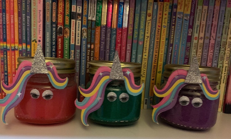 May include: Three glass jars decorated with unicorn faces. The jars are filled with pink, green, and purple glitter. The jars are sitting on a shelf in front of a bookshelf.