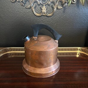 May include: A vintage copper tea kettle with a black handle and spout. The kettle sits on a dark wood tray with a gold-colored trim. The background includes a decorative mirror and a dark wall.