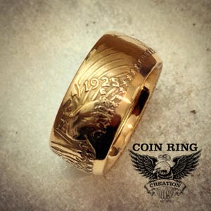 May include: A gold-colored coin ring with the year 1925 visible. The ring features detailed engravings of an eagle and other designs. The words "COIN RING" and a logo with an eagle are also visible.