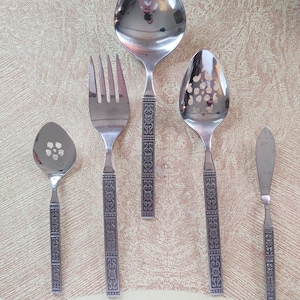 Puede incluir: Cinco utensilios de plata con diseños ornamentados en los mangos. Los utensilios incluyen una cuchara ranurada, un tenedor, una cuchara de servir, una cuchara ranurada pequeña y un cuchillo para mantequilla.