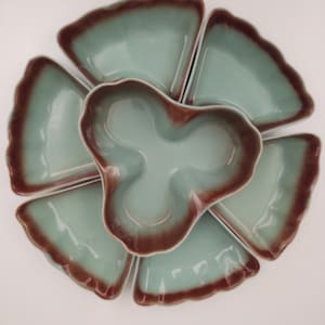 Puede incluir: Un juego de seis platos de servir de cerámica verde con bordes marrones. Los platos están dispuestos en forma de flor con un cuenco central.