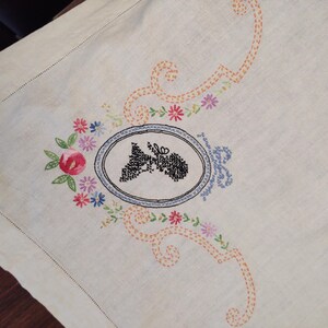 May include: White linen tablecloth with embroidered floral design and a black silhouette of a person's head in a blue oval frame.