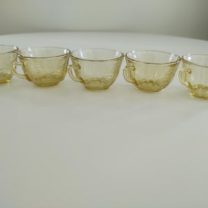 May include: Five vintage amber-colored glass teacups with handles. Each cup has a delicate floral pattern etched into the glass. The cups are arranged in a row on a white surface, showcasing their elegant design.