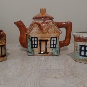 Può includere: Un set da tè in ceramica a tema cottage. La teiera, la lattiera e lo zuccheriera sono tutte a forma di piccole case con tetti marroni e bordi verdi. Ogni pezzo ha una finestra e una porta.