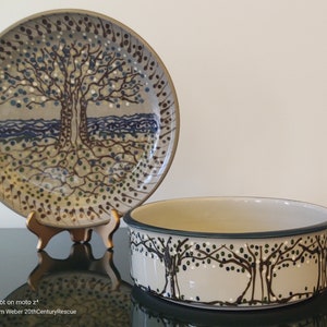 Könnte beinhalten: Ein Keramik-Teller- und Schüssel-Set mit einem Baumdesign. Der Teller, der auf einem Holzständer ausgestellt ist, zeigt einen Baum mit blauen, weißen und braunen Akzenten. Die Schüssel hat ein ähnliches Baummuster um ihr Äußeres, mit einem dunkelgrünen Rand.