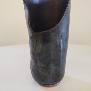 May include: A dark, handcrafted ceramic vase with a unique, asymmetrical shape. The vase has a dark, mottled finish with hints of green and brown. The rim curves outward, and the base is a lighter brown color.