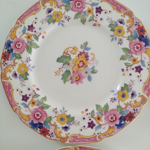Puede incluir: Platos de cerámica blancos con un diseño floral. Los platos tienen un borde rosa con un borde festoneado y están decorados con flores rosas, amarillas, azules y moradas. Un grupo de flores está en el centro.