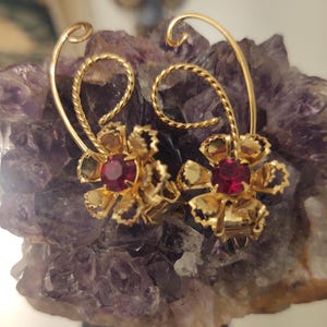May include: A pair of gold-toned floral earrings with a red gemstone center. The earrings have a curved stem design and are set against a purple, textured background.