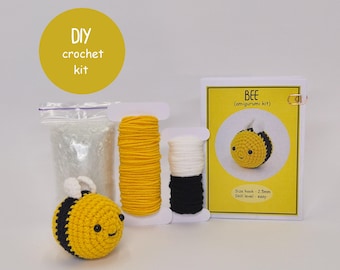 Peut inclure: Un kit de crochet DIY pour un amigurumi abeille. Le kit comprend du fil jaune, blanc et noir, de la bourre et un patron. L'abeille finie est jaune et noire avec un visage souriant. Le texte sur le kit dit "BEE (amigurumi kit)".