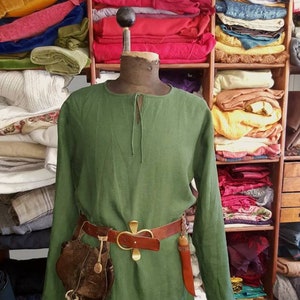 May include: A green linen tunic with long sleeves and a brown leather belt with a gold buckle. The tunic is on a mannequin and there are stacks of fabric in the background.