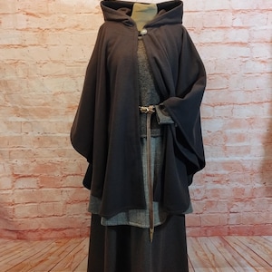 Könnte beinhalten: Ein dunkelgrauer Kapuzenmantel mit passendem langem Rock und einem grauen Tweedkleid. Der Umhang hat weite Ärmel und einen braunen Ledergürtel. Das Outfit wird vor einer Backsteinmauer und einem Holzboden präsentiert.