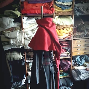 Könnte beinhalten: Ein roter Kapuzenumhang mit einem langen, fließenden Rock. Der Umhang ist über eine Schaufensterpuppe gelegt und von Regalen mit Stoff umgeben.