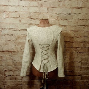 Peut inclure: Une chemise blanche à manches longues avec un dos à lacets et un motif floral. La chemise est sur un mannequin devant un mur de briques.