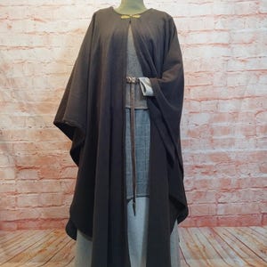 Pode incluir: Uma capa marrom escura com capuz e um fecho de metal decorativo. A capa é usada sobre uma saia xadrez cinza e branca.