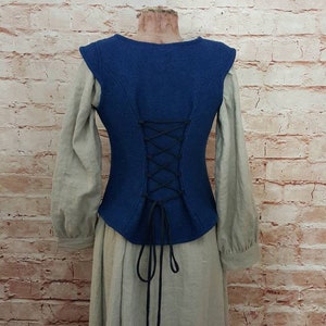 Könnte beinhalten: Eine blaue ärmellose Weste mit Schnürung im Rücken. Die Weste ist aus dickem Stoff gefertigt und hat eine figurbetonte Silhouette. Die Weste wird über einem langärmeligen weißen Hemd und einem langen beigefarbenen Rock getragen.