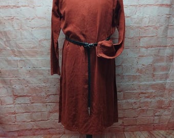 May include: A rust-colored long-sleeved dress with a black belt and a small pouch. The dress is displayed on a mannequin against a brick wall background. The text at the bottom of the image says, "The belt is not part of the offer."