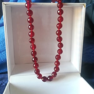 Op de afbeelding: Een ketting van gefacetteerde, dieprode kralen, gepresenteerd in een witte doos. De kralen zijn geregen aan een rood koord en hebben een edelsteenachtige uitstraling. De doos is open en onthult de ketting tegen een blauwe achtergrond.
