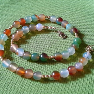 Può includere: Una collana di perline con una varietà di pietre preziose rotonde e colorate nelle tonalità dell'arancione, del verde, del blu e del grigio. Le perline sono infilate con piccole perline color argento e una chiusura argentata. La collana è esposta su una superficie verde.