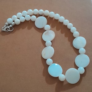 Può includere: Una collana bianca con perline rotonde e ovali di conchiglia. Ha una chiusura argentata ed è presentata su una superficie marrone. Il design combina piccole perline rotonde e perline ovali piatte più grandi.