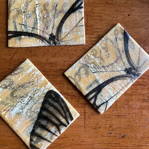 May include: Three square coasters with a yellow background and a black butterfly design. The butterflies are partially visible on each coaster.