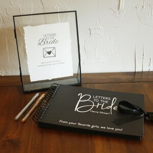 May include: Black scrapbook with a white ribbon and the text "Letters to the Bride" and "Hanna Stewart". The scrapbook is on a wooden table with two silver pens and a glass frame with a white sign that says "Letters to the Bride" and has a picture of an envelope with a heart.
