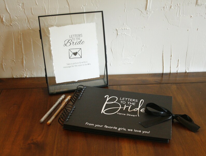 May include: Black scrapbook with a white ribbon and the text "Letters to the Bride" and "Hanna Stewart". The scrapbook is on a wooden table with two silver pens and a glass frame with a white sign that says "Letters to the Bride" and has a picture of an envelope with a heart.