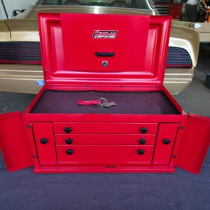 May include: A red Snap-on tool chest with the lid and side doors open, revealing three drawers and a key. The chest is on a dark surface, with a gold car in the background.