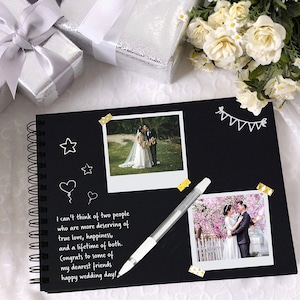 May include: A black scrapbook with a spiral binding, decorated with white washi tape, white stars, and a heart. The scrapbook has two photos of a wedding couple, one in a park and one in a garden. The text on the page reads "I can't think of two people who are more deserving of true love, happiness, and a lifetime of both. Congrats to some of my dearest friends happy wedding day!"