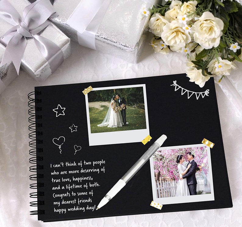 May include: A black scrapbook with a spiral binding, decorated with white washi tape, white stars, and a heart. The scrapbook has two photos of a wedding couple, one in a park and one in a garden. The text on the page reads "I can't think of two people who are more deserving of true love, happiness, and a lifetime of both. Congrats to some of my dearest friends happy wedding day!"