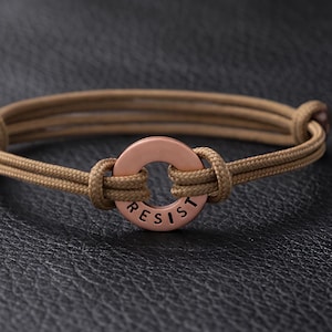 May include: A tan cord bracelet with a copper-colored circular centerpiece. The word "RESIST" is inscribed on the centerpiece. The bracelet is knotted and has a minimalist design.