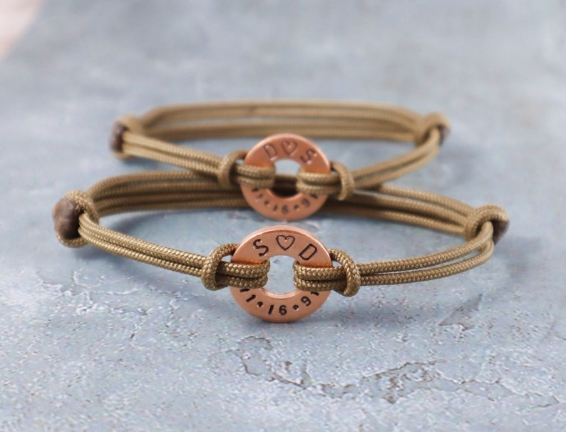 May include: Two olive green paracord bracelets with copper round charms. The charms have the letters "S & D" and "D & S" with a heart symbol between the letters and a date engraved on the bottom.