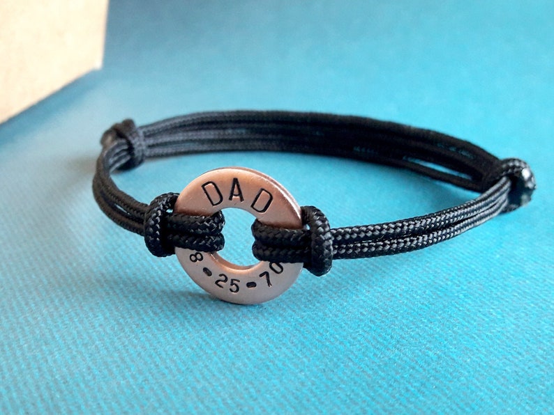 May include: A black cord bracelet with a copper-colored round charm that says "DAD 8-25-70".