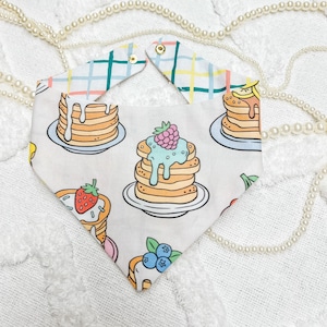 May include: A white bandana with a checkered pattern on the top and a pattern of colorful pancakes with toppings on the bottom.