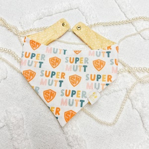 May include: A white bandana with a repeating pattern of the words "Super Mutt" in various colors and a small logo of a dog wearing a cape. The bandana has two snaps on the top for fastening.