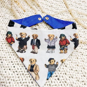May include: White dog bandana with a blue striped collar. The bandana features a repeating pattern of cartoon teddy bears dressed in various outfits, including suits, sweaters, and athletic wear. The bandana is secured with two small wooden buttons.