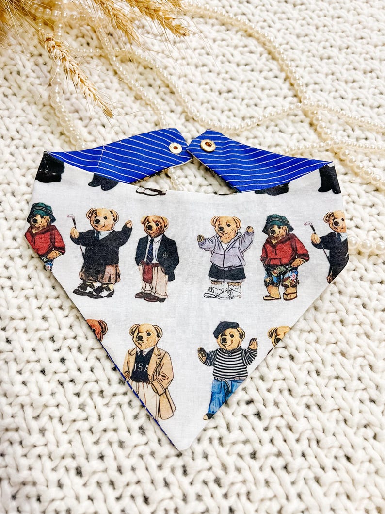 May include: A white dog bandana with a blue striped collar. The bandana displays a pattern of cartoon teddy bears in different outfits, such as suits and sportswear. The bandana is fastened with two small wooden buttons. The measurements are in centimeters.