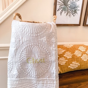 May include: A white, quilted fabric basket with a decorative design and the embroidered word "Ghost" in green. The basket has woven handles and is placed on a wooden surface. A decorative pillow and framed artwork are in the background.