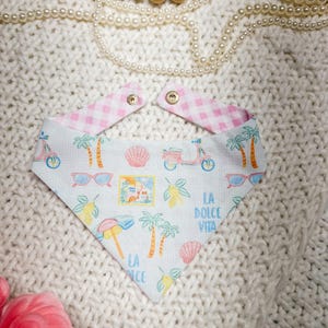 May include: A white bandana with a pink gingham pattern on the neck straps. The bandana features a colorful print of scooters, palm trees, sunglasses, lemons, and the words "La Dolce Vita" and "La Nice".