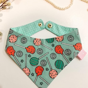 May include: A green bandana with a white polka dot pattern and a print of red, green, and pink cake pops on sticks. The bandana has two snap closures at the top.
