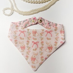 May include: A pink and white pet bandana featuring a bunny and floral design. The bandana has a pink bow and flower pattern. The top of the bandana has a pink floral pattern and gold snap closures. A pearl necklace is in the background.