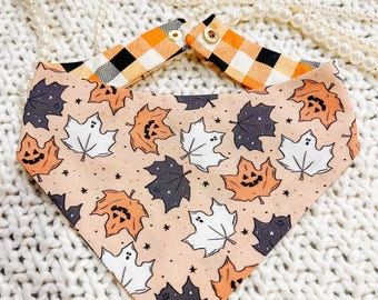 May include: A Halloween-themed dog bandana with a peach background and a pattern of orange jack-o'-lantern leaves, white ghost leaves, and dark gray leaves. The bandana's neck strap is orange and white checkered.