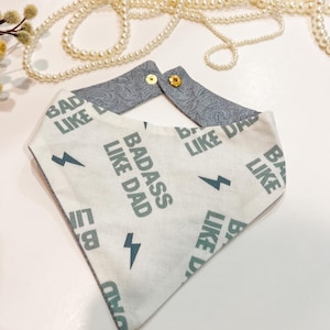 May include: A white bandana with a green and white pattern. The bandana has the text "Badass Like Dad" repeated on it. The bandana is folded in a triangle shape and has a grey paisley fabric on the back.