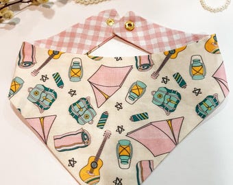 May include: A pink and white bandana with a camping theme. The bandana features a pattern of tents, sleeping bags, lanterns, guitars, and backpacks. The bandana has a pink and white gingham trim.