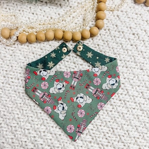 May include: A green dog bandana with a festive pattern featuring white dogs, nutcrackers, and candy canes. The bandana has a green snowflake pattern on the adjustable neck straps. The background includes decorative beads and a white knitted surface.