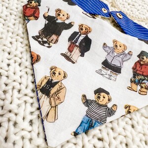 May include: Dog bandana featuring a pattern of teddy bears in different outfits, such as a blazer and golf attire, on a white background. The reverse side is blue with white stripes. It has gold press studs.
