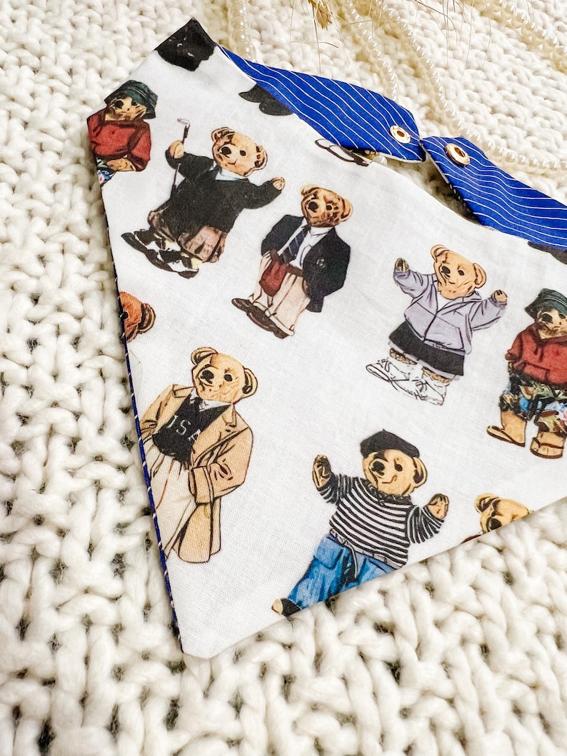May include: Dog bandana featuring a pattern of teddy bears in different outfits, such as a blazer and golf attire, on a white background. The reverse side is blue with white stripes. It has gold press studs.