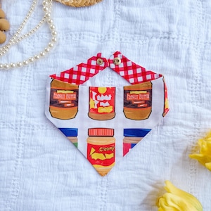 May include: A triangular pet bandana featuring a white background with a repeating pattern of peanut butter jars. The fabric includes jars with red, blue, and brown lids and labels reading Peanut Butter and Creamy. The top edge has a red and white gingham trim.