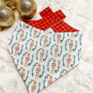 May include: A white bandana with a red and white checkered pattern on the back. The front of the bandana features a repeating pattern of nutcracker figures and green holly sprigs.