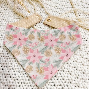May include: A dog bandana with a festive floral pattern. The bandana features pink poinsettias, green leaves, and gold pinecones on a white background. The top portion is tan with white polka dots and gold snaps.