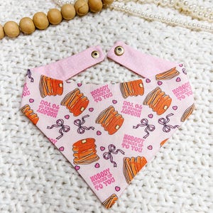 May include: Pink dog bandana with a pancake pattern and the text "NOBODY STACKS UP TO YOU". The bandana has a pink polka dot trim and is secured with snaps. The design includes orange pancakes, bows, and hearts.